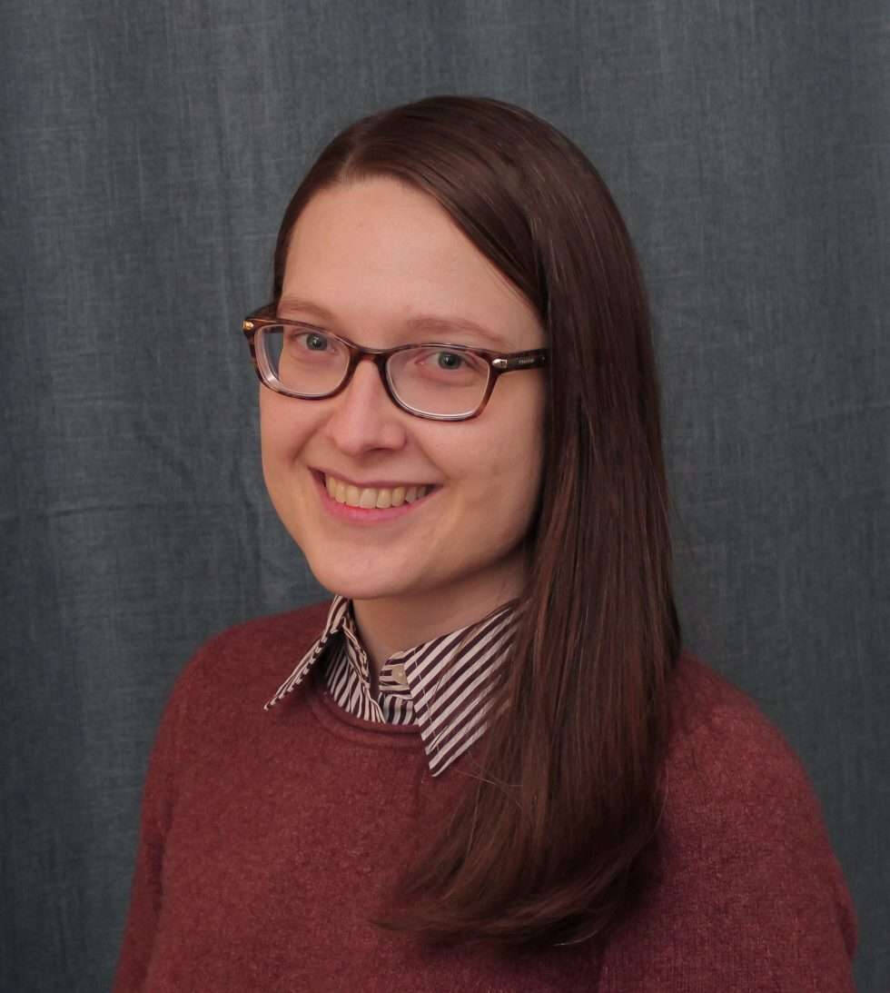 A person with straight, medium-length brown hair is wearing a maroon sweater over a black and white striped collared shirt. The background consists of a gray fabric curtain.